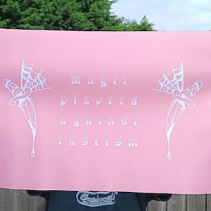 Puede incluir: Póster rosa con texto e ilustraciones en blanco. El texto dice "magic players against fascism". Las ilustraciones son dagas estilizadas con telarañas. El póster está sostenido por una persona.