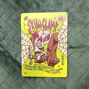 May include: A trading card with a neon green background features a skull clamped between two brown wooden planks. The card reads "SKULLCLAMP" in pink letters and includes the text "EQUIPPED CREATURE GETS -1/-1. WHEN EQUIPPED CREATURE DIES, DRAW 2 CARDS. EQUIP 1".