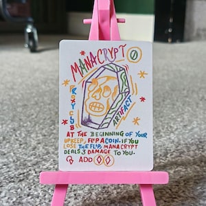 Puede incluir: Una tarjeta blanca con el texto "MANA CRYPT" y "ARTIFACT" en letras coloridas, con una ilustración de calavera. La tarjeta se muestra en un caballete rosa. El texto en la parte inferior describe las reglas del juego.