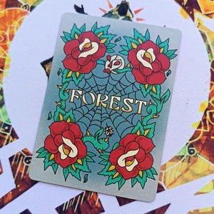 May include: A rectangular card featuring a tattoo-style design. Four red roses with yellow centers and green leaves surround a blue spiderweb. The word "FOREST" is in gold letters across the centre.