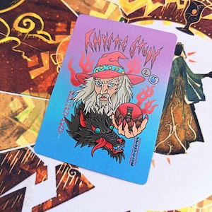 Könnte beinhalten: Eine rechteckige Karte mit einem blau-violetten Farbverlauf. Die Karte zeigt einen Zauberer mit rotem Hut, der eine rote Kugel mit einem Turm hält. Unter dem Zauberer befindet sich ein schwarzer Wolf mit roten Flammen. Die Karte lautet "Mystic Story".