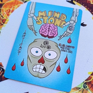 Könnte beinhalten: Eine blaue Karte mit den Worten "MIND STONE" in Gelb, mit einem Cartoon-Gehirn, einem Schädel mit dem Wort "ADD" und dem Text "SACRIFICE MIND STONE: DRAW A CARD". Rote Blutstropfen sind ebenfalls vorhanden.
