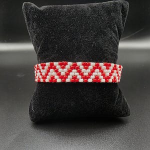 May include: A beaded bracelet with a repeating pattern of red and white beads. The bracelet is displayed on a black velvet cushion against a black background. The beads are small and tightly woven together.