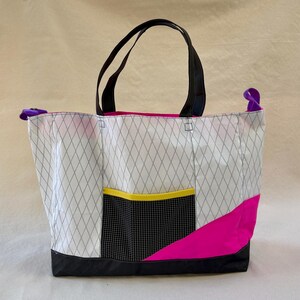 May include: A tote bag featuring a white diamond-patterned sailcloth body, black handles, and a black base. It includes a black grid-patterned front pocket with a yellow trim, a vibrant pink diagonal accent panel, and purple side attachment loops.