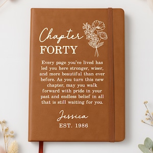 May include: A tan leather-bound journal featuring a white floral line drawing and the text "Chapter FORTY. Every page you've lived has led you here stronger, wiser, and more beautiful than ever before. As you turn this new chapter, may you walk forward with pride in your past and endless belief in all that is still waiting for you. Jessica EST. 1986."