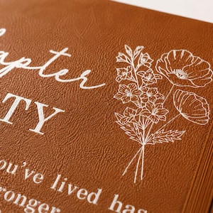 May include: A brown leather-textured journal cover featuring white printed text that reads Chapter FORTY and a white line-art illustration of a floral bouquet. The cover includes a vertical elastic band on the right side for closure.