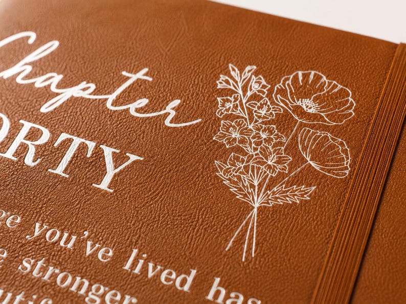 May include: A brown leather-textured journal cover featuring white printed text that reads Chapter FORTY and a white line-art illustration of a floral bouquet. The cover includes a vertical elastic band on the right side for closure.