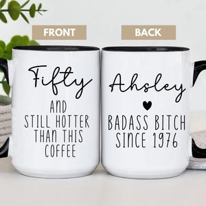 May include: White ceramic coffee mug with a black handle and rim. The front of the mug reads "Fifty AND STILL HOTTER THAN THIS COFFEE." The back says "Ahsley BADASS BITCH SINCE 1976" with a heart symbol.