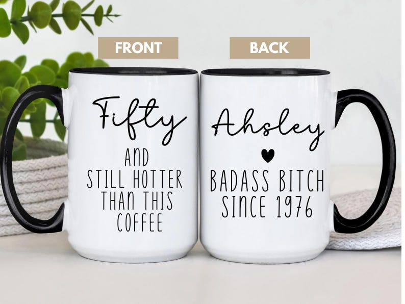 May include: White ceramic coffee mug with a black handle and rim. The front of the mug reads "Fifty AND STILL HOTTER THAN THIS COFFEE." The back says "Ahsley BADASS BITCH SINCE 1976" with a heart symbol.