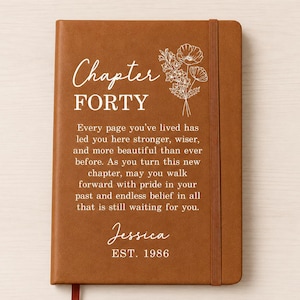 May include: A tan notebook with an elastic closure featuring white text that reads Chapter FORTY, followed by an inspirational quote, the name Jessica, and EST. 1986. A white line art illustration of a floral bouquet is positioned above the text.