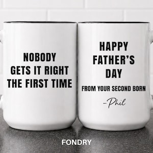 May include: Two white ceramic mugs with black handles and rims. One mug reads "NOBODY GETS IT RIGHT THE FIRST TIME" in black text. The other mug says "HAPPY FATHER'S DAY FROM YOUR SECOND BORN -Phil" in black text.