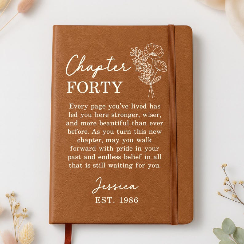 May include: A tan leather-bound journal featuring a white floral line drawing and the text "Chapter FORTY. Every page you've lived has led you here stronger, wiser, and more beautiful than ever before. As you turn this new chapter, may you walk forward with pride in your past and endless belief in all that is still waiting for you. Jessica EST. 1986."