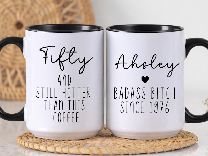 May include: Two white ceramic coffee mugs with black handles and rims. One mug reads "Fifty AND STILL HOTTER THAN THIS COFFEE." The other mug says "Ahsley BADASS BITCH SINCE 1976" with a heart symbol. Both mugs are on a woven coaster.