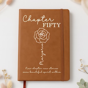 May include: A brown leather journal with the words "Chapter FIFTY" and "Marjorie" in white script. A white rose graphic is centered on the cover. The journal also has the quote "New chapter new stories, same beautiful spirit within."