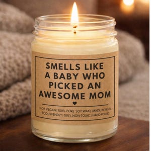 May include: A lit candle in a clear glass jar with a brown label. The label reads "Smells like a baby who picked an awesome mom." The candle is 9 oz and made with vegan soy wax. The candle is hand poured.