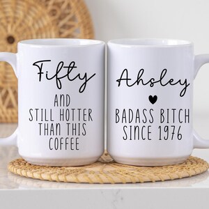 May include: Two white ceramic coffee mugs with black text. One mug says "Fifty AND STILL HOTTER THAN THIS COFFEE." The other says "Ahsley BADASS BITCH SINCE 1976" with a black heart. Both mugs have large handles.