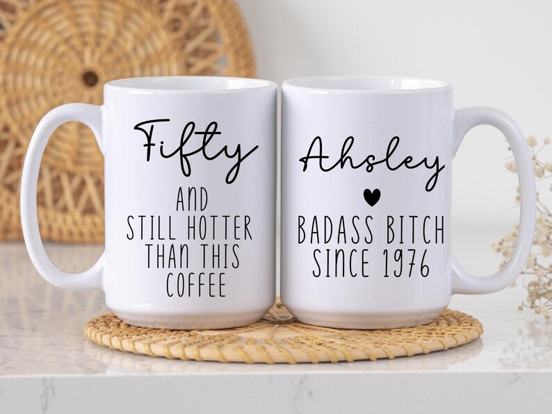 May include: Two white ceramic coffee mugs with black text. One mug says "Fifty AND STILL HOTTER THAN THIS COFFEE." The other says "Ahsley BADASS BITCH SINCE 1976" with a black heart. Both mugs have large handles.