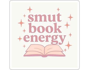 May include: A square sticker with the words "smut book energy" in pink, surrounded by orange starbursts. An open book illustration is at the bottom, also in pink, on a cream background.