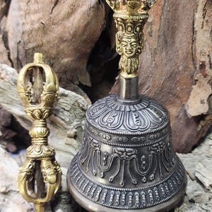 May include: Two ornate brass and silver-toned ritual objects. A bell with detailed patterns and a small, hand-held vajra. The bell has a detailed face at the top. The objects are on a wood background.
