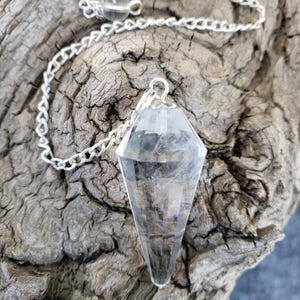 May include: A clear crystal pendulum with a silver chain and a small crystal bead at the top. The diamond-shaped pendulum hangs against a textured, weathered wooden surface.