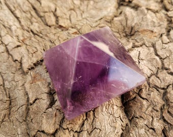 May include: A faceted amethyst pyramid crystal, showcasing shades of purple and violet. The crystal is set against a textured, light brown wooden surface. This decorative crystal is a unique home decor item.