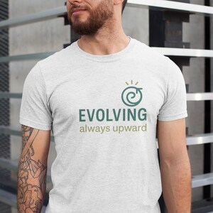 Puede incluir: Una camiseta gris claro con las palabras "EVOLVING always upward" en verde. La camiseta presenta un diseño de espiral verde con un pequeño sol encima. La camiseta es de manga corta.