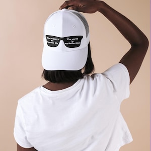 Peut inclure: Casquette de camionneur blanche avec un motif de lunettes de soleil noir et blanc. Le design des lunettes de soleil comprend le texte "What triggers Me Teaches Me" sur le verre gauche et "The world is My Reflection" sur le verre droit.