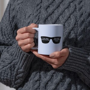 May include: A white ceramic mug with a black graphic of sunglasses and the text "I see what needs healing. The outer reflects the inner." The mug is held by a person wearing a gray cable knit sweater. The mug has a white handle.