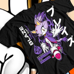 Puede incluir: Camiseta negra con un gráfico de Blaze the Cat, un personaje de la serie Sonic the Hedgehog. El diseño incluye al personaje en morado y blanco, con texto japonés y las palabras "Blaze the Cat" en blanco.