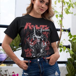 May include: Black t-shirt featuring a graphic of a character with a horned mask and the text "Who Laughs" in red. The design has a comic book style with red accents. The person is wearing a denim skirt.