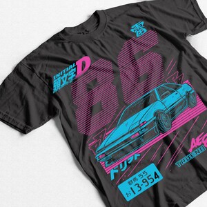 Puede incluir: Camiseta negra con un diseño gráfico azul y rosa de un coche y el número "86". La camiseta incluye texto japonés y un diseño de matrícula. El diseño general tiene un tema de carreras retro.