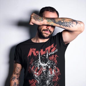 May include: Black t-shirt featuring a graphic of a skeletal figure in a horned helmet with red accents and Japanese text. The words "WHO LAUGHS" are also visible. The shirt is worn by a person with tattoos on their arms.