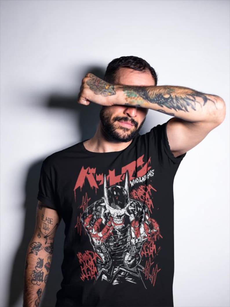 May include: Black t-shirt featuring a graphic of a skeletal figure in a horned helmet with red accents and Japanese text. The words "WHO LAUGHS" are also visible. The shirt is worn by a person with tattoos on their arms.