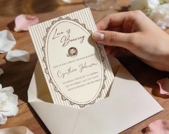 Può includere: Un invito per un bridal shower con il testo "Love is Brewing" in una cornice ovale ornata. L'invito è in una busta color crema, con accenti floreali e un bicchiere di champagne su una superficie di legno.