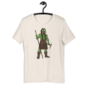 May include: Cream-coloured t-shirt featuring a cartoon illustration of a green frog warrior holding a spear and a small axe. The frog is wearing a brown leather skirt and boots. The shirt is hanging on a black hanger.