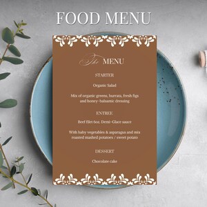 Puede incluir: Una tarjeta de menú marrón con borde floral blanco y texto "FOOD MENU" en la parte superior. El menú enumera opciones de entrantes, platos principales y postres, incluyendo ensalada orgánica, filete de ternera y pastel de chocolate. La tarjeta descansa sobre un plato azul.