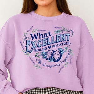 Op de afbeelding: Een lichtpaarse sweatshirt met de tekst "What Excellent Boiled Potatoes" in een bloemmotief. De afbeelding bevat twee aardappelillustraties en de woorden "An Exemplary Vegetable". De sweatshirt heeft lange mouwen en een ronde hals.