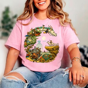 Puede incluir: Camiseta rosa claro con una ilustraci&oacute;n colorida de un drag&oacute;n, personajes y un paisaje. El dise&ntilde;o incluye un drag&oacute;n, figuras y una luna llena. La camiseta est&aacute; hecha de un material suave.