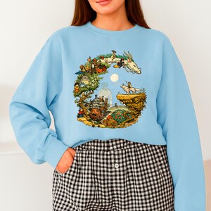 Puede incluir: Sudadera azul claro con una ilustraci&oacute;n colorida de un drag&oacute;n, personajes y un paisaje. El gr&aacute;fico es un dise&ntilde;o detallado y caprichoso. La sudadera tiene mangas largas y cuello redondo.