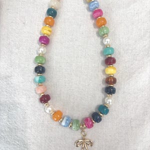 Puede incluir: Un collar que presenta una variedad de cuentas multicolores, lisas y redondeadas en tonos rosa, azul, amarillo, verde, naranja y blanco, intercaladas con pequeñas cuentas espaciadoras de metal dorado y un dije de lazo de metal dorado.