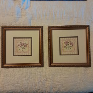 May include: Two square wall art pieces featuring ornate bronze-colored frames with decorative beading. Each print displays a floral illustration centered on a cream-colored mat, with one showing purple lilies and the other showing pink roses.