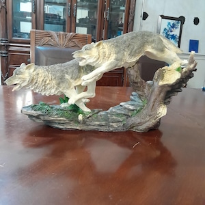 May include: A detailed figurine featuring two grey and white wolves in a dynamic running pose. The wolves are positioned on a textured base that resembles a rocky, mossy landscape with a prominent tree trunk element, capturing a wild scene.