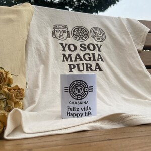 May include: Cream-coloured t-shirt with the text "YO SOY MAGIA PURA" and three stylized faces. A white mug with the text "CHASKINA Feliz vida Happy life" is in front of the shirt. A beige pillow is on the left.