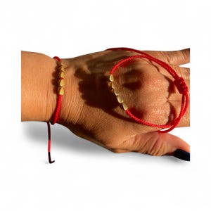 May include: Two red string bracelets with gold-colored cube beads. One bracelet is tied on a wrist, and the other is held in a hand. The bracelets are displayed against a white background.