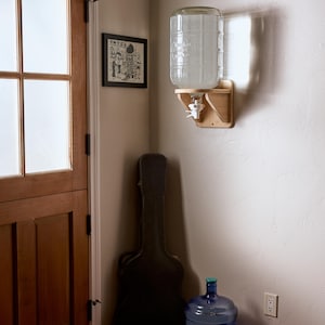 May include: A clear glass water dispenser with a wooden base and spigot is mounted on a wall. A large blue water jug sits on the floor. A framed picture and a guitar case are also in the corner.