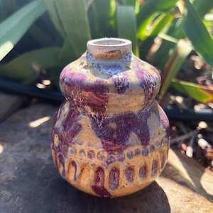 May include: A handcrafted ceramic vase with a bulbous shape and a narrow neck. The vase features a two-tone design with a tan base and a deep burgundy pattern. The top rim is a light tan color. The vase is sitting on a dark stone surface.
