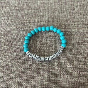 May include: A turquoise beaded bracelet with the word "TRANSFORMED" spelled out in white letter beads. The bracelet is made of round, smooth beads and is displayed on a textured brown surface.