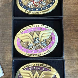 May include: Three oval-shaped, gold-colored metal belt buckles in black boxes. Each buckle has the text "BE HUMBLE BE STRONG" and "BE BRAVE BE BADASS". The buckles feature the USN logo and various designs.