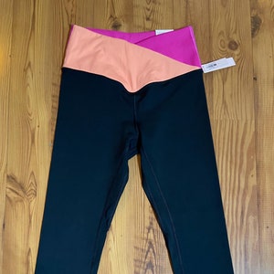 Puede incluir: Leggings negros con una cintura rosa y melocotón. La cintura tiene un diseño diagonal.