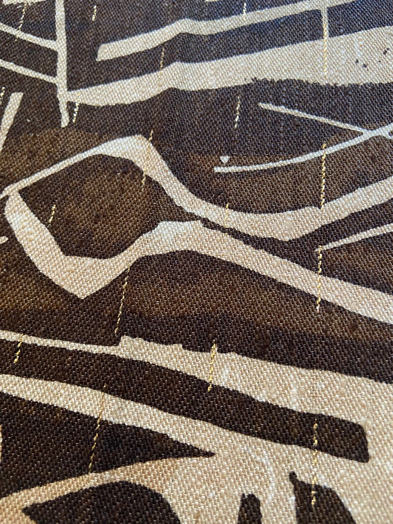 Pu&ograve; includere: Primo piano di un tessuto strutturato con un motivo ripetuto. Il design presenta forme marrone scuro e bianco sporco con sottili fili d'oro che attraversano la trama. Il tessuto ha un aspetto tessuto.